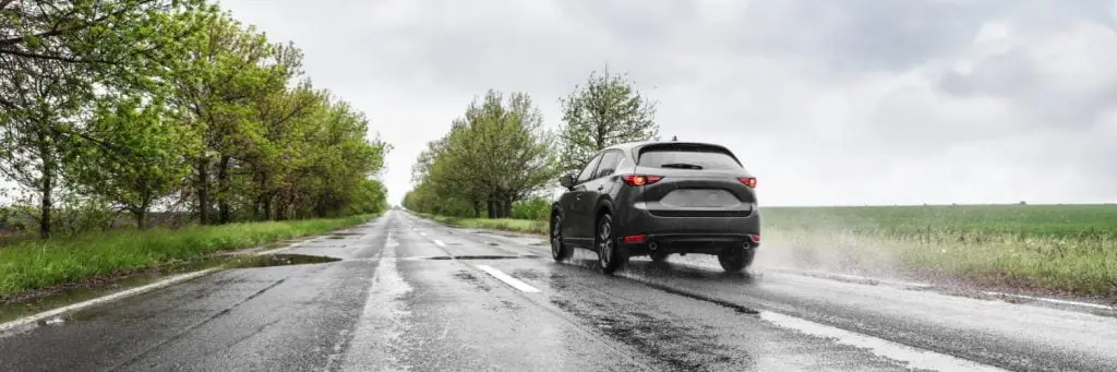 A black car drives down a wet, empty road on a rainy day. The road is flanked by green fields and trees, and the sky is overcast with clouds. Superior Tire: Tire Shop and Auto Repair Near Me | Barrie | Danforth | Downsview | Markham | Mississauga | Oak Ridges | Scarborough | Thornhill | Toronto | https://superiortire.ca/