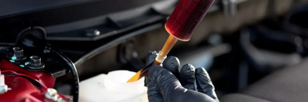 A person wearing black gloves uses a syringe to extract brake fluid from a white reservoir under a car hood. The fluid is a transparent brownish-red color, hinting at maintenance or inspection work being done. Superior Tire: Tire Shop and Auto Repair Near Me | Barrie | Danforth | Downsview | Markham | Mississauga | Oak Ridges | Scarborough | Thornhill | Toronto | https://superiortire.ca/