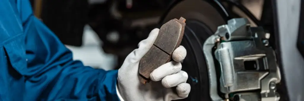Mechanic in blue overalls and white gloves holding a worn car brake pad in front of a brake disc. The scene is set in a workshop, highlighting the importance of brake maintenance for vehicle safety. Superior Tire: Tire Shop and Auto Repair Near Me | Barrie | Danforth | Downsview | Markham | Mississauga | Oak Ridges | Scarborough | Thornhill | Toronto | https://superiortire.ca/