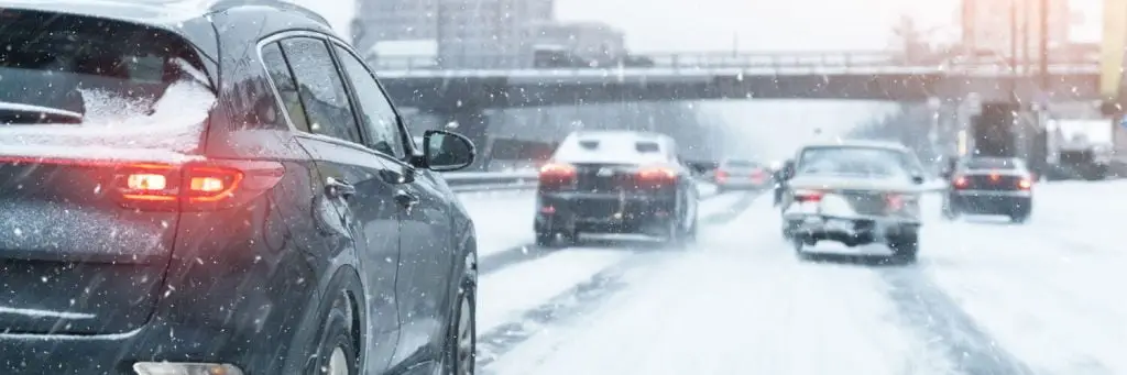 Cars driving cautiously on a snow-covered highway. Snow is falling, and vehicles have their lights on. An overpass can be seen in the background under a cloudy sky. Superior Tire: Tire Shop and Auto Repair Near Me | Barrie | Danforth | Downsview | Markham | Mississauga | Oak Ridges | Scarborough | Thornhill | Toronto | https://superiortire.ca/