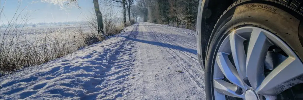 A close-up of a car tire on a snow-covered rural road, with a row of bare trees lining the path. The sky is clear and blue, and sunlight casts long shadows across the snow. Superior Tire: Tire Shop and Auto Repair Near Me | Barrie | Danforth | Downsview | Markham | Mississauga | Oak Ridges | Scarborough | Thornhill | Toronto | https://superiortire.ca/