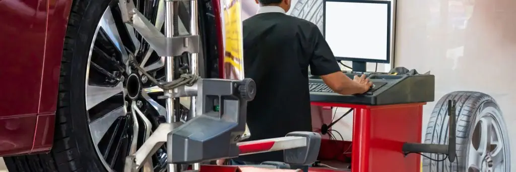 A mechanic in a black shirt works at a computer next to a red car on a lift, focusing on wheel alignment tools. Two tire images are on the wall behind him. Superior Tire: Tire Shop and Auto Repair Near Me | Barrie | Danforth | Downsview | Markham | Mississauga | Oak Ridges | Scarborough | Thornhill | Toronto | https://superiortire.ca/