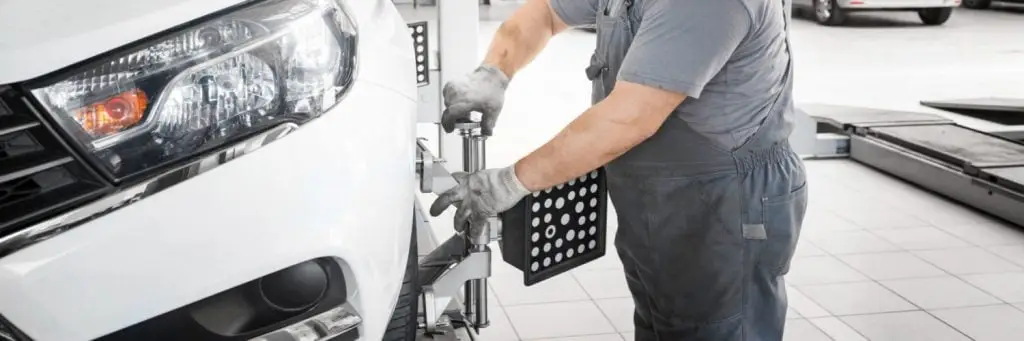 A mechanic in gray overalls and gloves adjusts equipment on the wheel of a white car inside an auto repair shop. The focus is on aligning the vehicle with specialized tools, in a garage with tiled flooring. Superior Tire: Tire Shop and Auto Repair Near Me | Barrie | Danforth | Downsview | Markham | Mississauga | Oak Ridges | Scarborough | Thornhill | Toronto | https://superiortire.ca/