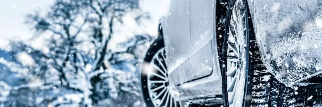 Close-up of a car tire on a snowy road with snowflakes falling. The background shows blurred trees covered in snow, highlighting the winter scene. The focus is on the wheel and tire, emphasizing traction in icy conditions. Superior Tire: Tire Shop and Auto Repair Near Me | Barrie | Danforth | Downsview | Markham | Mississauga | Oak Ridges | Scarborough | Thornhill | Toronto | https://superiortire.ca/