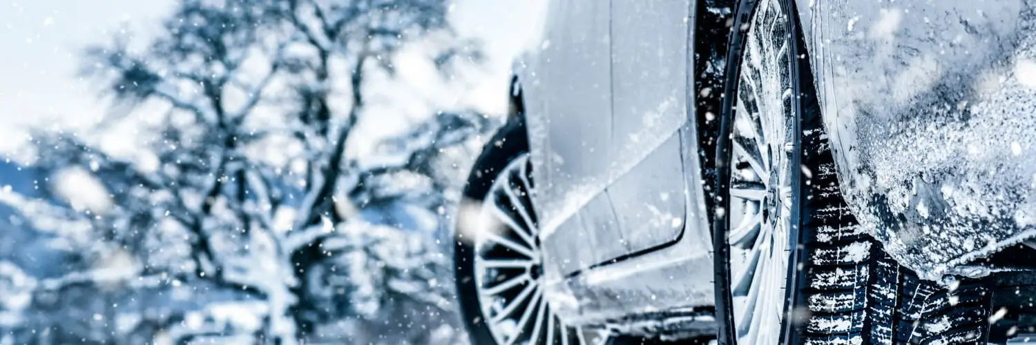 Close-up of a car tire on a snowy road with snowflakes falling. The background shows blurred trees covered in snow, highlighting the winter scene. The focus is on the wheel and tire, emphasizing traction in icy conditions. Superior Tire: Tire Shop and Auto Repair Near Me | Barrie | Danforth | Downsview | Markham | Mississauga | Oak Ridges | Scarborough | Thornhill | Toronto | https://superiortire.ca/