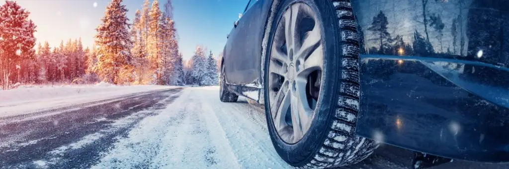 Close-up of a cars rear tire on a snowy road. The landscape features snow-covered trees under a bright blue sky. The sunlight creates a warm, glowing effect on the trees and road. Superior Tire: Tire Shop and Auto Repair Near Me | Barrie | Danforth | Downsview | Markham | Mississauga | Oak Ridges | Scarborough | Thornhill | Toronto | https://superiortire.ca/