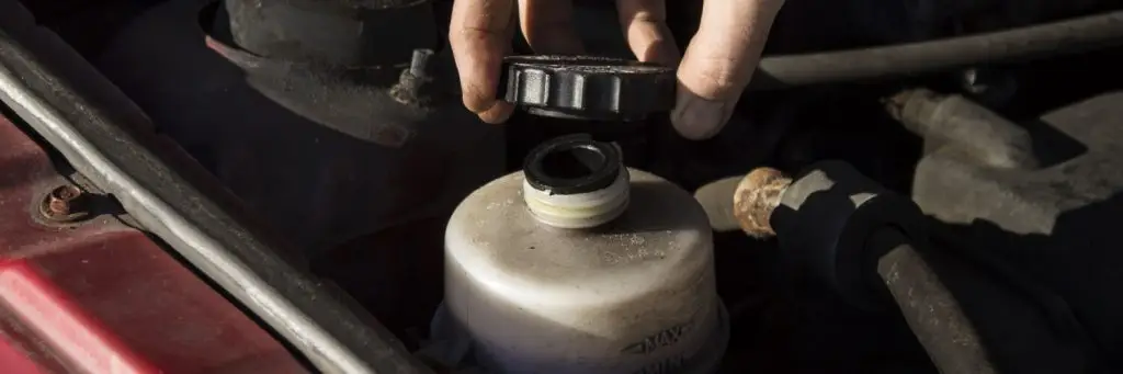 A hand is unscrewing the cap of a vehicles brake fluid reservoir under the hood. The reservoir is translucent with a black cap, and various engine components are visible in the background. Superior Tire: Tire Shop and Auto Repair Near Me | Barrie | Danforth | Downsview | Markham | Mississauga | Oak Ridges | Scarborough | Thornhill | Toronto | https://superiortire.ca/