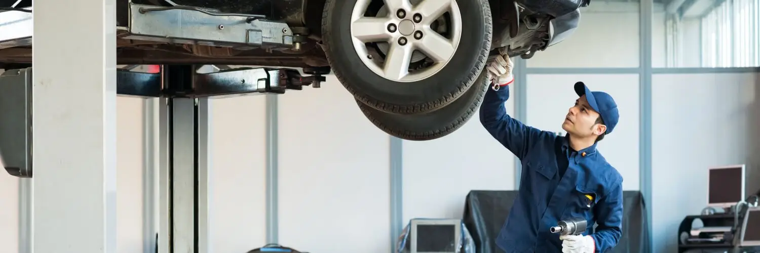 A mechanic in a blue uniform and cap inspects the underside of a raised car in a garage. He holds a tool in one hand, focusing on the vehicles components. The workshop is equipped with various tools and equipment. Superior Tire: Tire Shop and Auto Repair Near Me | Barrie | Danforth | Downsview | Markham | Mississauga | Oak Ridges | Scarborough | Thornhill | Toronto | https://superiortire.ca/
