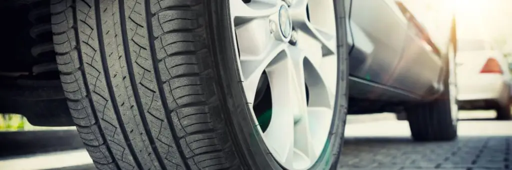 Close-up of a car tire on a paved surface, with another car visible in the background. The sun shines brightly, casting shadows and creating a warm glow on the vehicles. The tires tread pattern is clearly visible. Superior Tire: Tire Shop and Auto Repair Near Me | Barrie | Danforth | Downsview | Markham | Mississauga | Oak Ridges | Scarborough | Thornhill | Toronto | https://superiortire.ca/