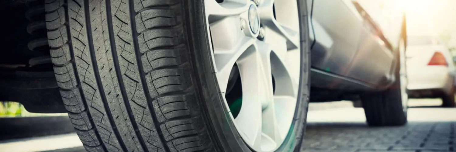 Close-up of a car tire on a paved surface, with another car visible in the background. The sun shines brightly, casting shadows and creating a warm glow on the vehicles. The tires tread pattern is clearly visible. Superior Tire: Tire Shop and Auto Repair Near Me | Barrie | Danforth | Downsview | Markham | Mississauga | Oak Ridges | Scarborough | Thornhill | Toronto | https://superiortire.ca/