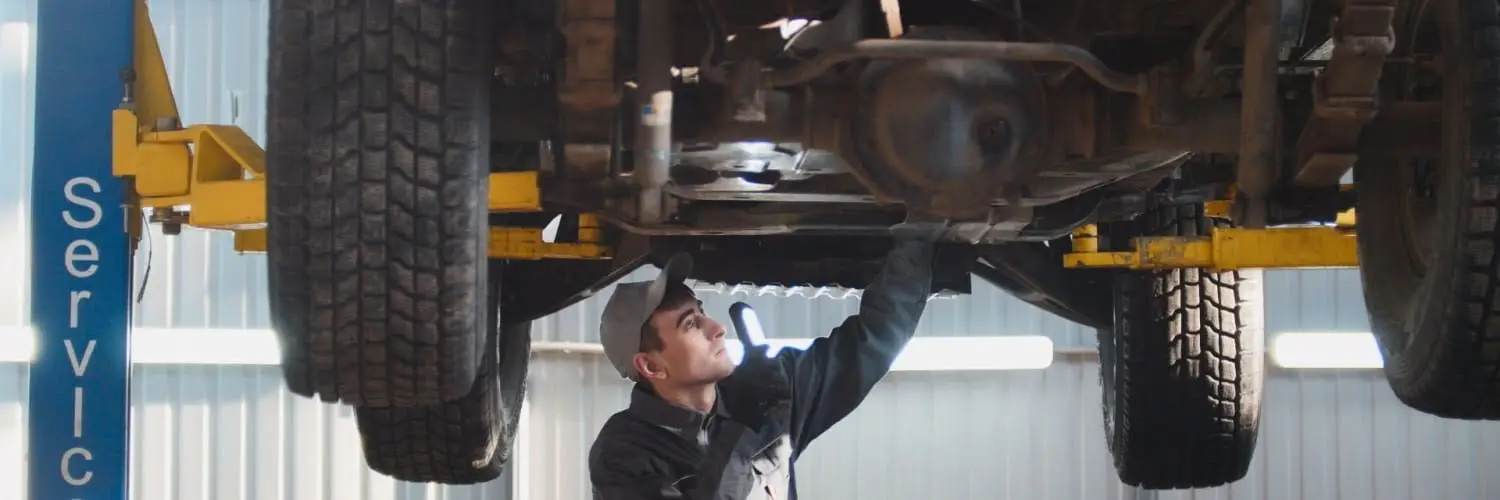 Mechanic in a cap inspects the underside of a raised truck on a lift in a garage. He uses a flashlight to check components. The lift is labeled Servic and the garage is well-lit, with windows and corrugated walls visible in the background. Superior Tire: Tire Shop and Auto Repair Near Me | Barrie | Danforth | Downsview | Markham | Mississauga | Oak Ridges | Scarborough | Thornhill | Toronto | https://superiortire.ca/