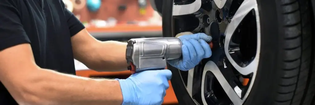 A person wearing blue gloves uses a power tool to tighten bolts on a car wheel in a garage. The focus is on their hands and the wheel, with a blurred background of tools and equipment. Superior Tire: Tire Shop and Auto Repair Near Me | Barrie | Danforth | Downsview | Markham | Mississauga | Oak Ridges | Scarborough | Thornhill | Toronto | https://superiortire.ca/