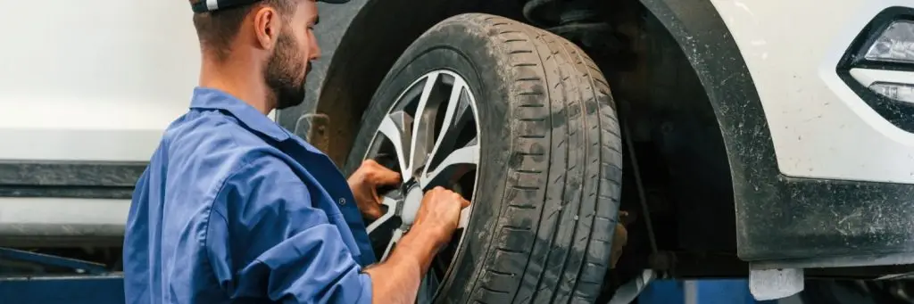 Mechanic in blue overalls and cap working on a white car elevated on a lift, adjusting the front tire. The setting appears to be an auto repair shop. Superior Tire: Tire Shop and Auto Repair Near Me | Barrie | Danforth | Downsview | Markham | Mississauga | Oak Ridges | Scarborough | Thornhill | Toronto | https://superiortire.ca/