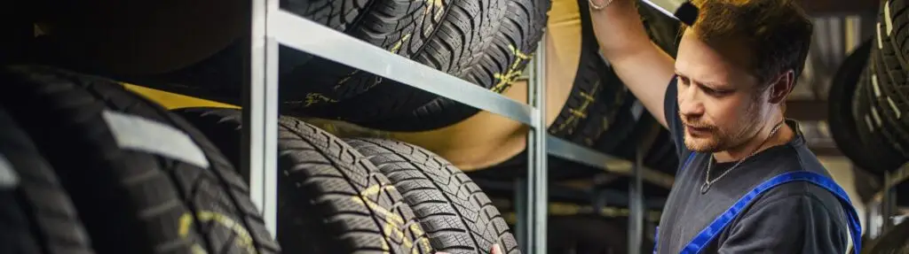 In a dimly lit industrial setting, a man in a workshop uniform meticulously examines tires on metal shelves. Surrounded by stacked rows of black tires, he focuses intently on his task, ensuring everything is ready for anyone searching for a tire change near me. Superior Tire: Tire Shop and Auto Repair Near Me | Barrie | Danforth | Downsview | Markham | Mississauga | Oak Ridges | Scarborough | Thornhill | Toronto | https://superiortire.ca/